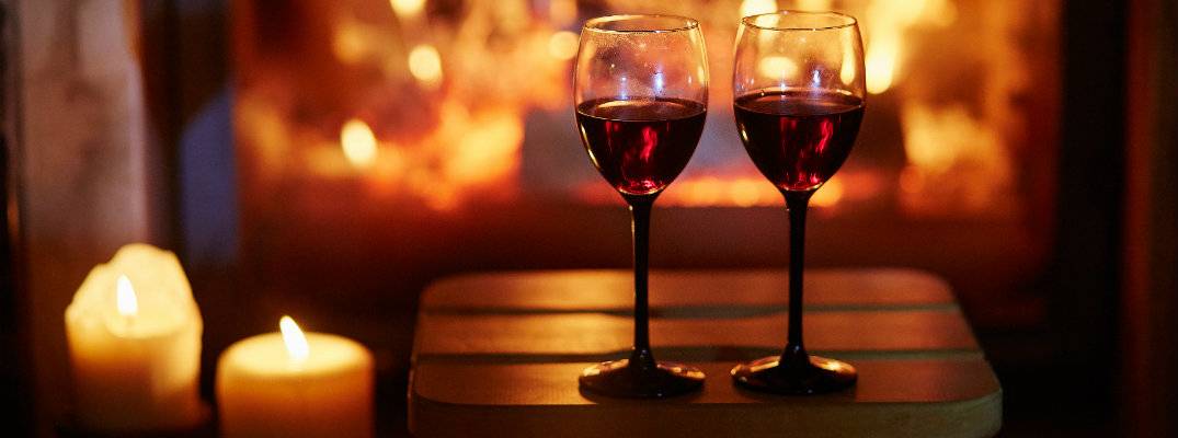 a couple of glasses of red wine set on a stool near lit candles and a fireplace b