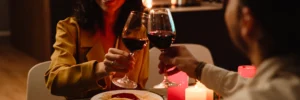 Casal em um primeiro encontro, brindando com taças de vinho tinto em uma mesa de jantar à luz de velas, com um prato de espaguete ao molho vermelho à frente.